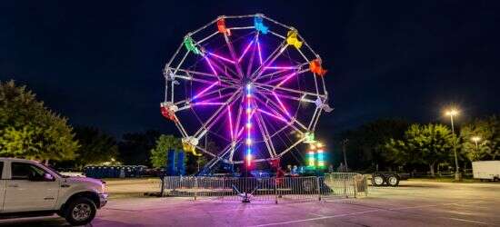 Colorful illuminated Ferris wheel rental attraction glowing at night with vibrant lights and a fenced seating area outdoors.