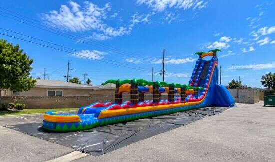 Large tropical inflatable water slide rental with colorful lanes and palm tree decorations set up outdoors on a sunny day.