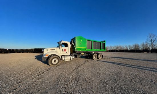Green roll-off dumpster truck parked in an open gravel yard