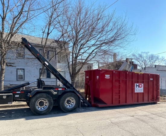A roll-off truck placing a red Huntley Disposal dumpster on a paved street in front of a residential house with bare trees.