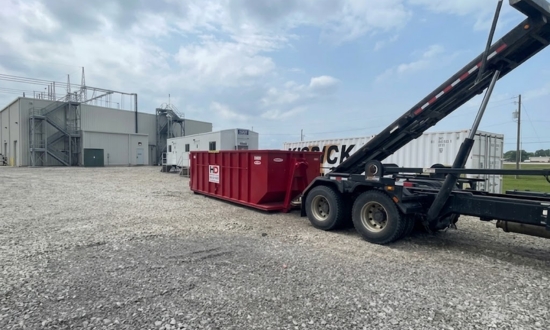 A roll-off dumpster truck unloading a large red Huntley Disposal dumpster onto a gravel industrial lot near commercial buildings.