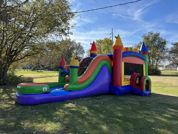 A colorful inflatable bounce house with bright red, yellow, green, and blue sections and towering castle-like spires stands on a grassy area in a park. The bounce house features a slide and a basketball hoop, set against a backdrop of trees and a clear blue sky.