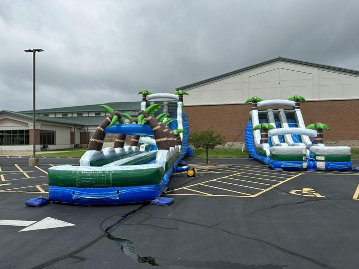 Two large inflatable water slides with a tropical theme, featuring palm tree designs and brown, blue, and green colors, are set up in an empty parking lot. The overcast sky above and the school building in the background suggest a cool or cloudy day.
