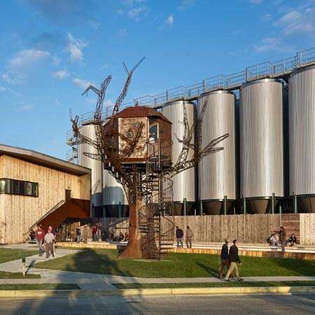 People walking near the Dogfish Head Brewery in Milton, Delaware, with its iconic steampunk treehouse and large silver fermentation tanks in the background.