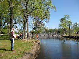 People fishing along the tree-lined banks of a calm river near a small dam on a clear day.
