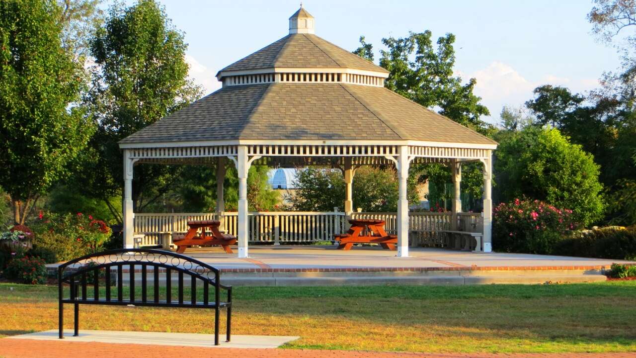 White gazebo with a shingled roof surrounded by greenery and picnic tables, located in a serene park setting in Milford, Delaware.