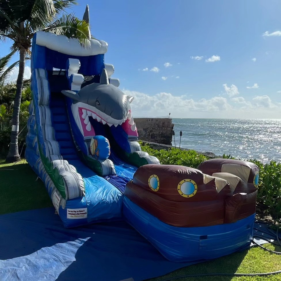 Shark-themed inflatable water slide with a giant smiling shark head on top and a pirate ship design at the base, positioned by the ocean under a clear blue sky, great for beach-themed party rentals in Oahu, HI, available at Oahu Jump.