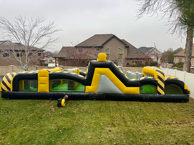 A large inflatable obstacle course rental set up on a grassy field at a community event. It is Yellow, green and black and ready for an adventurous day of play and competition