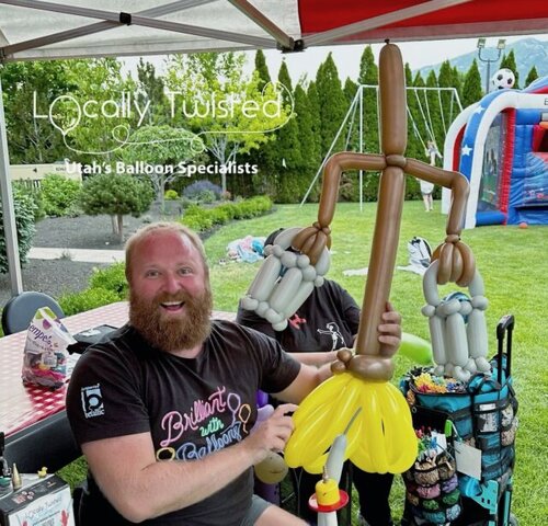 A man holding Lumière made out of balloons at a backyard party with green grass and trees behind him.
