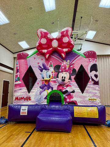  A girl joyfully poses in front of a colorful Minnie Mouse bounce house, set up indoors, ready for fun and play