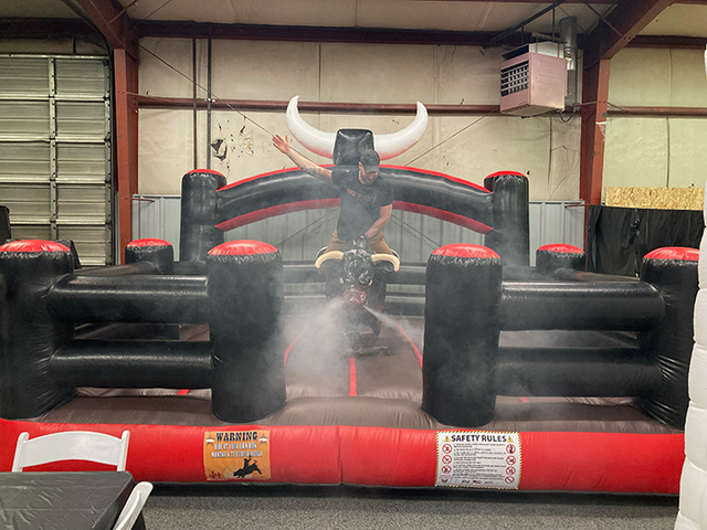 A mechanical bull set up on inflatable bed with a man riding it like a cowboy. The bull has steam coming out of its nose.