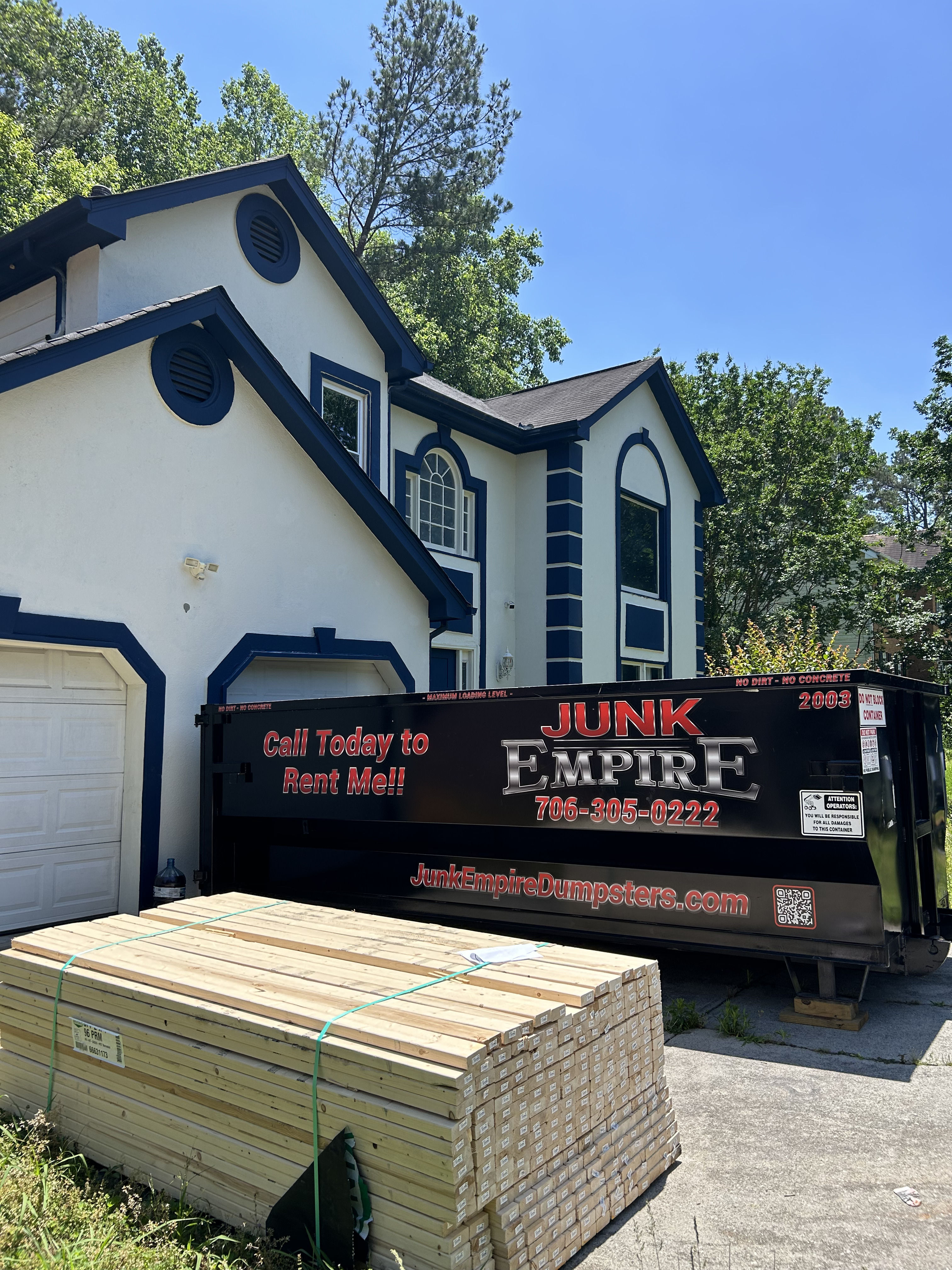 Junk Empire black dumpster container in residential driveway with lumber stack nearby