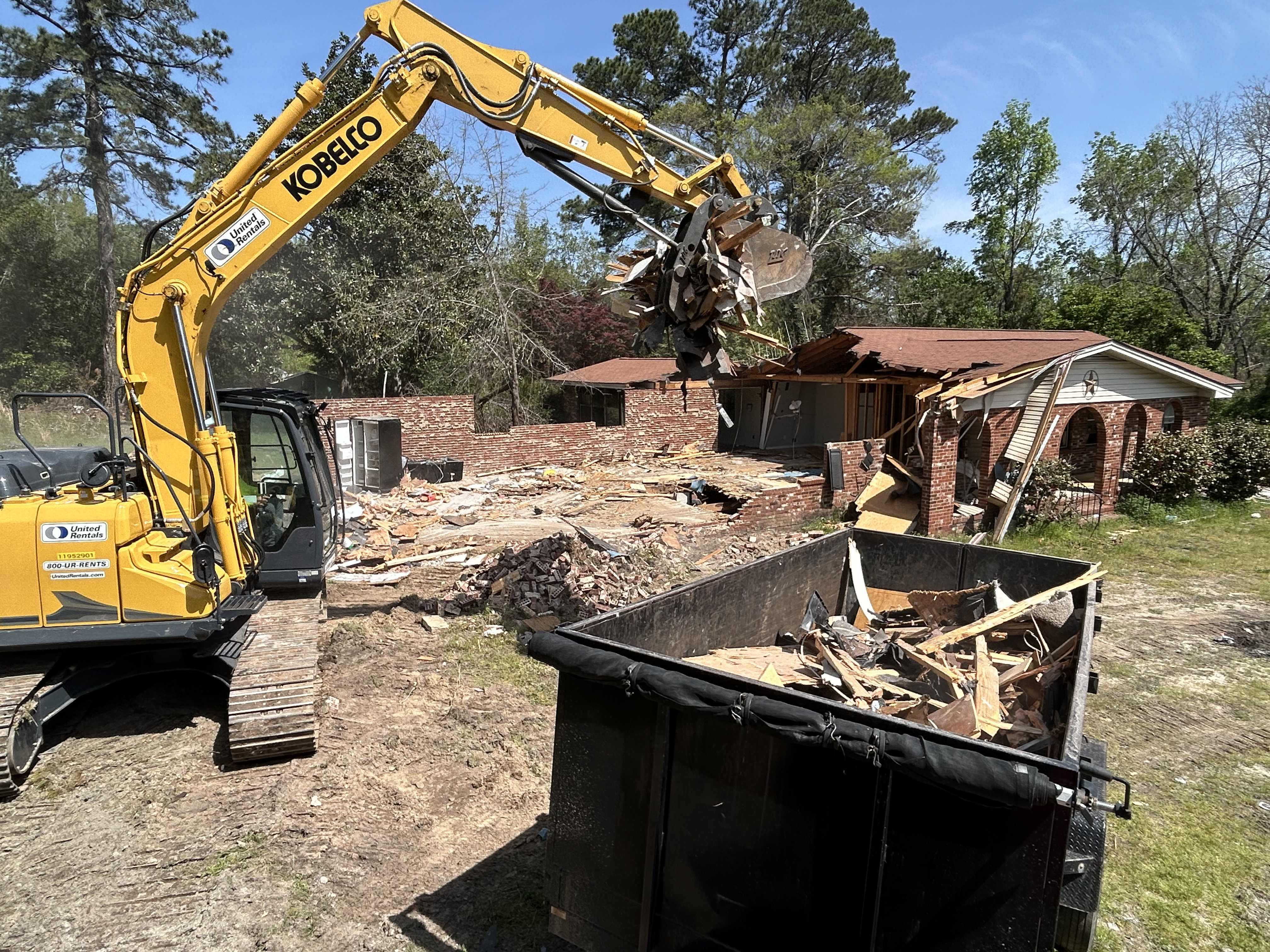 Junk Empire yellow excavator demolishing brick house with debris in black dumpster