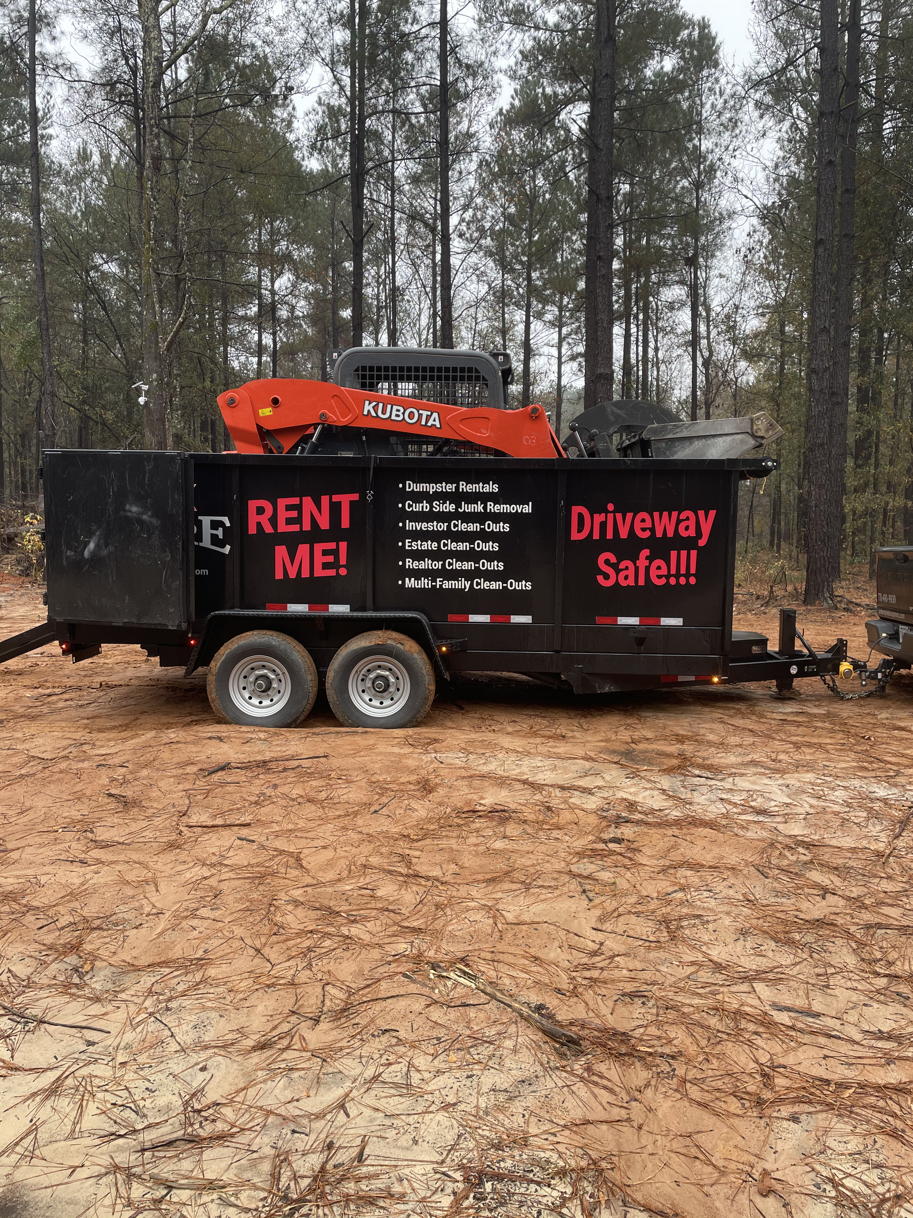 Junk Empire black dumpster with bold red RENT ME! and Driveway Safe!!! text, loaded with a Kubota machine for junk removal and clean-out services.