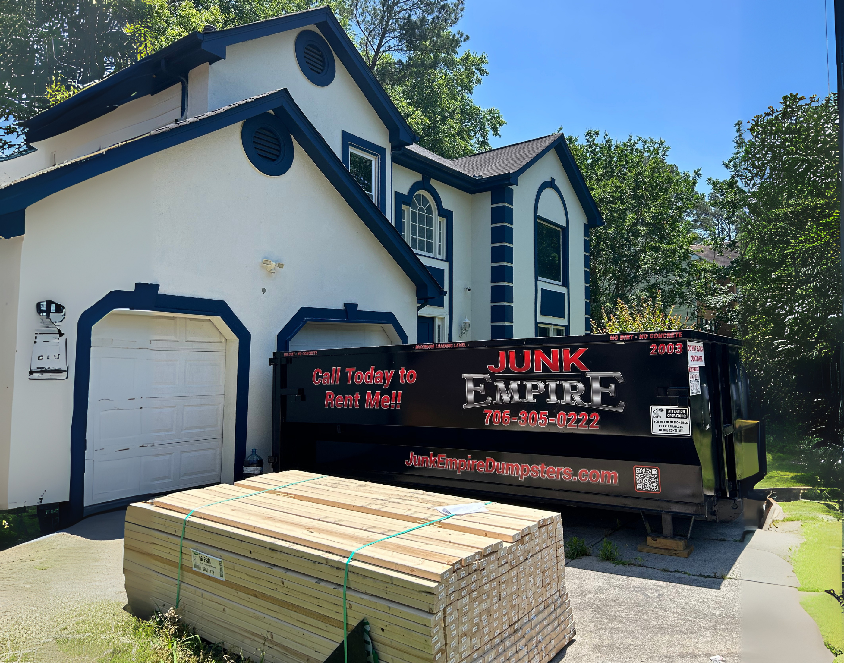 Junk Empire black dumpster container in residential driveway with lumber stack nearby