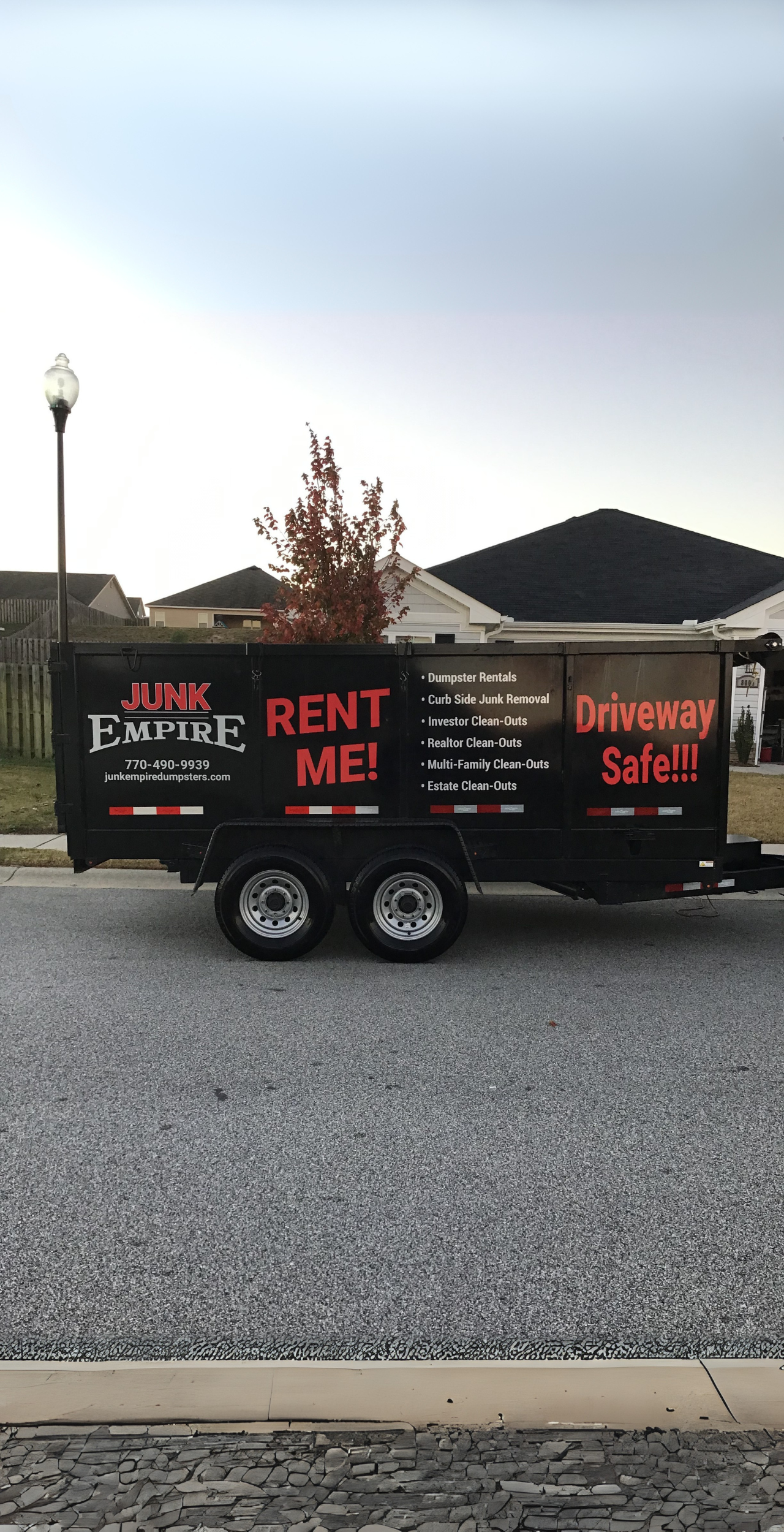 Junk Empire black dumpster rental trailer parked on residential street, featuring company branding and services such as junk removal, clean-outs, and driveway-safe rentals.