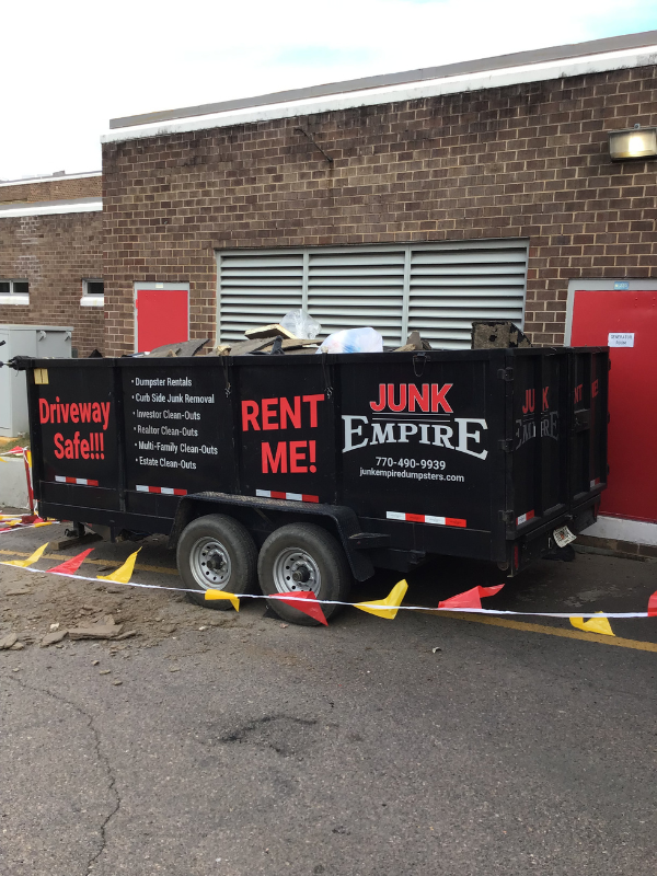 Junk Empire black dumpster trailer filled with debris, placed beside a brick building.