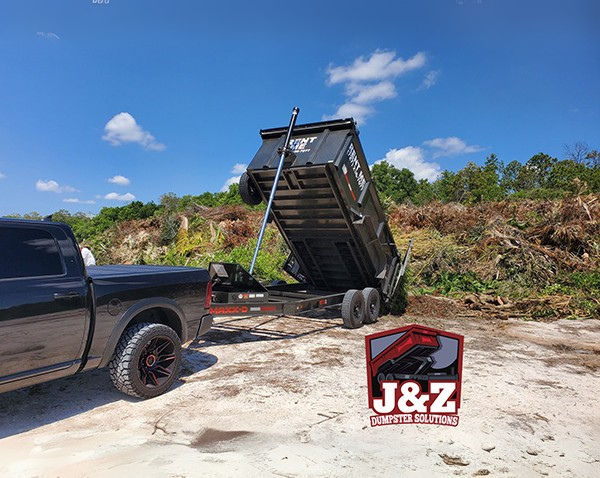 Black J&Z Dumpster Solutions dump trailer unloading debris at a land clearing site. Black J&Z Dumpster Solutions dump trailer unloading debris at a land clearing site.
