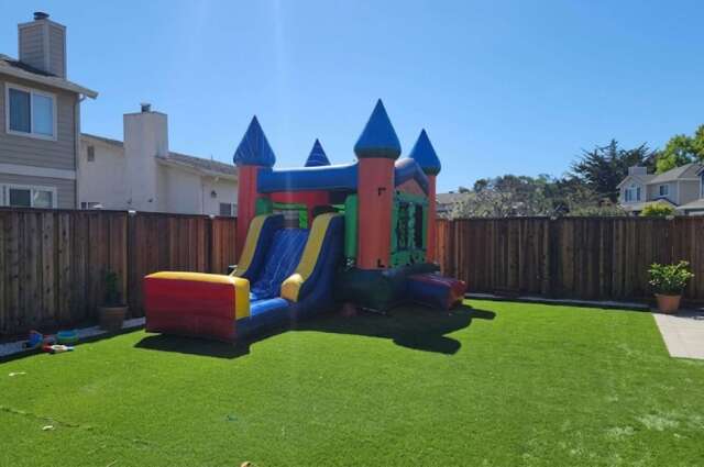 A blue and yellow inflatable bounce house and slide combo with castle turrets on an artificial grass lawn beside a wooden fence