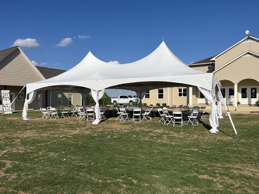 A white frame tent pitched on a lawn, set with round tables, folding chairs and string lights