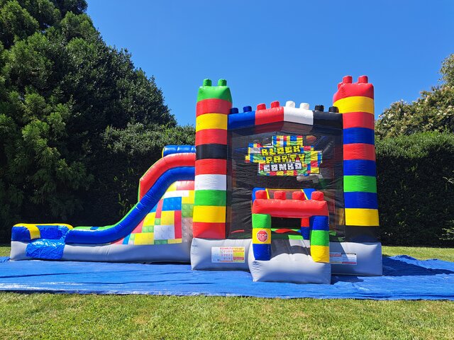 Colorful Lego-themed inflatable bounce house with attached slide set up on a blue tarp outdoors.