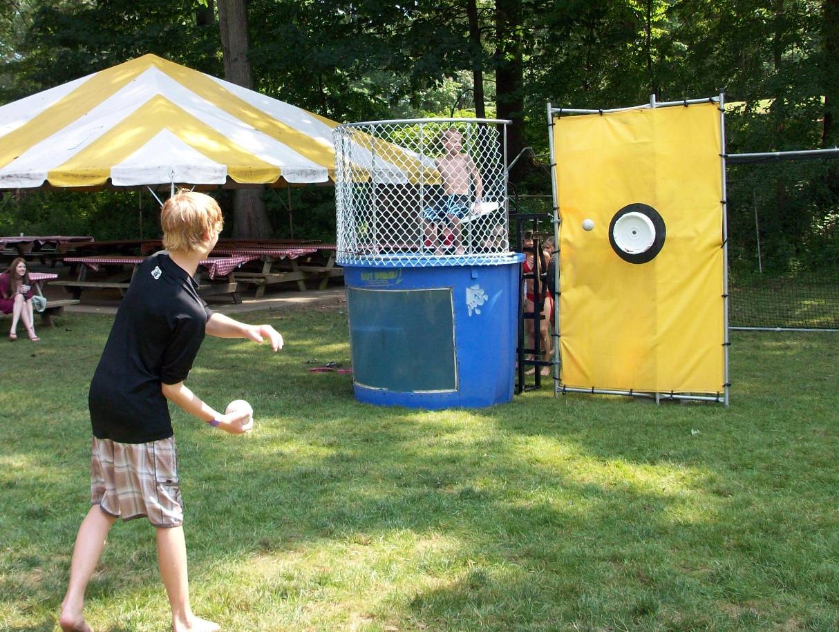 This dunk tank rental in Travelers Rest, SC is set up on grass at an outdoor park-style event with a striped tent and picnic tables nearby. A participant throws a ball at the bright yellow target, aiming to drop the seated guest into the water below. Ideal for school field days, church gatherings, birthday parties, corporate picnics, and community festivals throughout Travelers Rest, a dunk tank adds interactive fun and friendly competition to any outdoor celebration.
