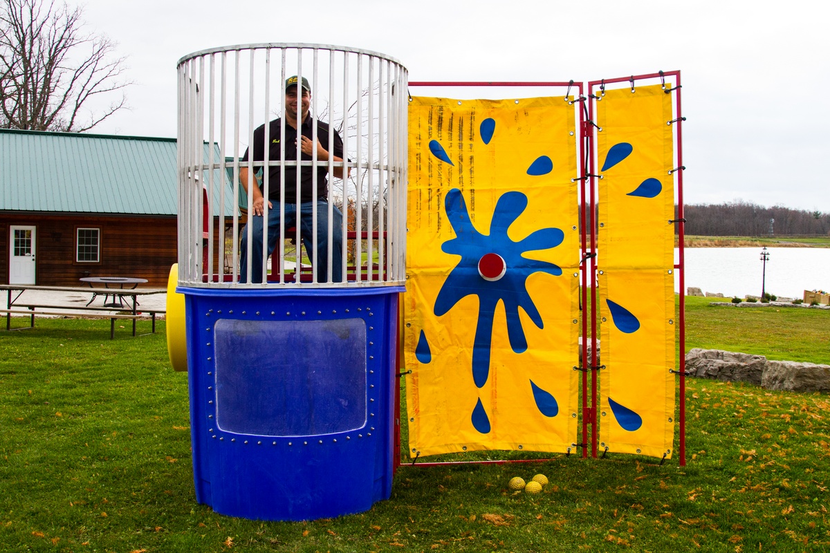 This dunk tank rental in Travelers Rest, SC is set up outdoors on grass near a pavilion and waterfront area, making it ideal for park events and community gatherings. The blue tank features a clear viewing window and enclosed safety cage, paired with a bright yellow splash-style target backdrop for high visibility. Perfect for school field days, church festivals, corporate picnics, and fundraisers throughout Travelers Rest, this dunk tank delivers interactive fun and memorable crowd engagement.