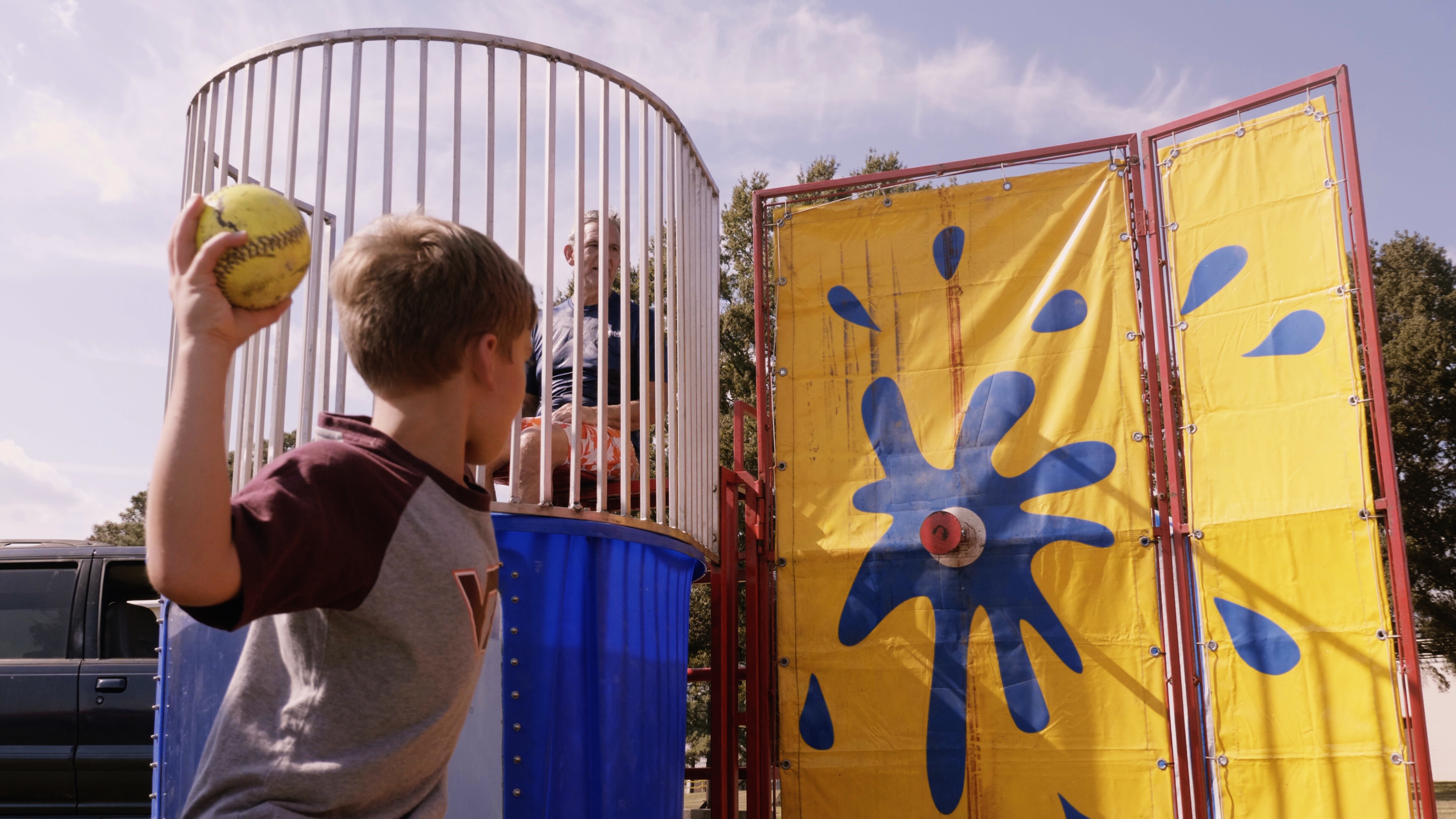 This dunk tank rental in Taylors, SC is set up outdoors for a community event, featuring a bright yellow splash-style target and enclosed safety cage. A child throws a softball at the target while a participant waits above the water tank. Perfect for school field days, church fundraisers, birthday parties, and corporate events in Taylors, this classic carnival attraction creates interactive fun and crowd engagement for all ages.
