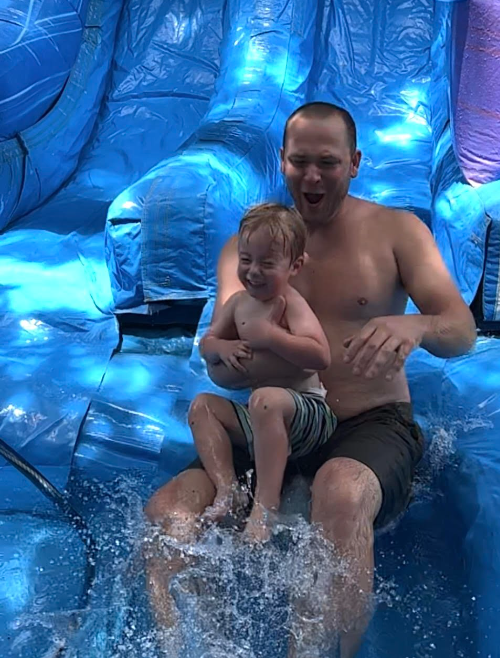 Father and son laughing together as they splash down a blue inflatable water slide during a summer rental in Berea, SC.