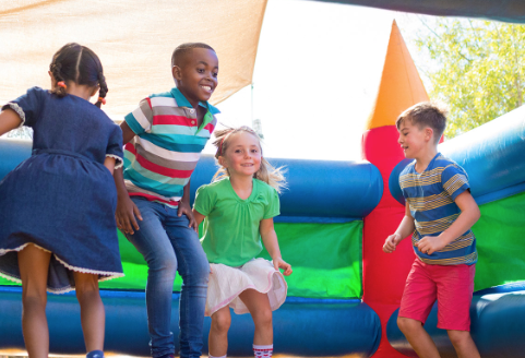 Four children laughing and jumping together inside a colorful inflatable bounce house under a sunshade on a bright day.