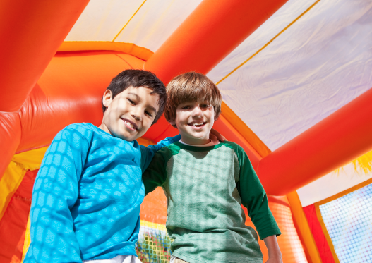 Two smiling boys standing inside a bright orange and yellow bounce house, with sunlight casting patterns through the mesh walls.