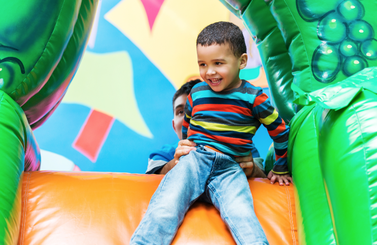 Smiling young boy in a striped shirt sliding down an inflatable slide with the help of an adult, surrounded by colorful inflatable walls.