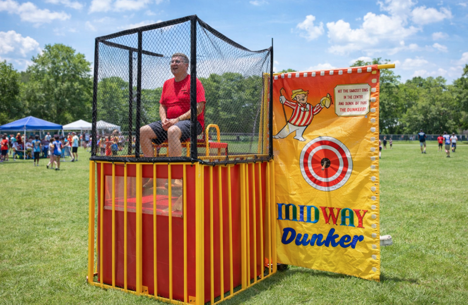 Yellow carnival dunk tank rental at a school field day in Fountain Inn, SC with adult participant on the seat and event crowd in the background.