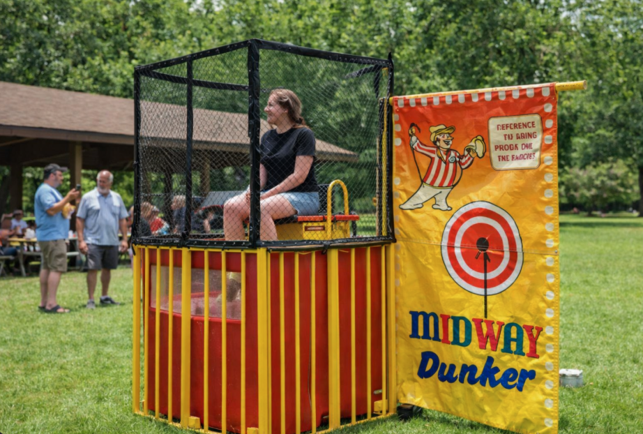 Yellow carnival dunk tank rental at a company picnic in Fountain Inn, SC with adult participant seated above the tank in a sunny park setting.
