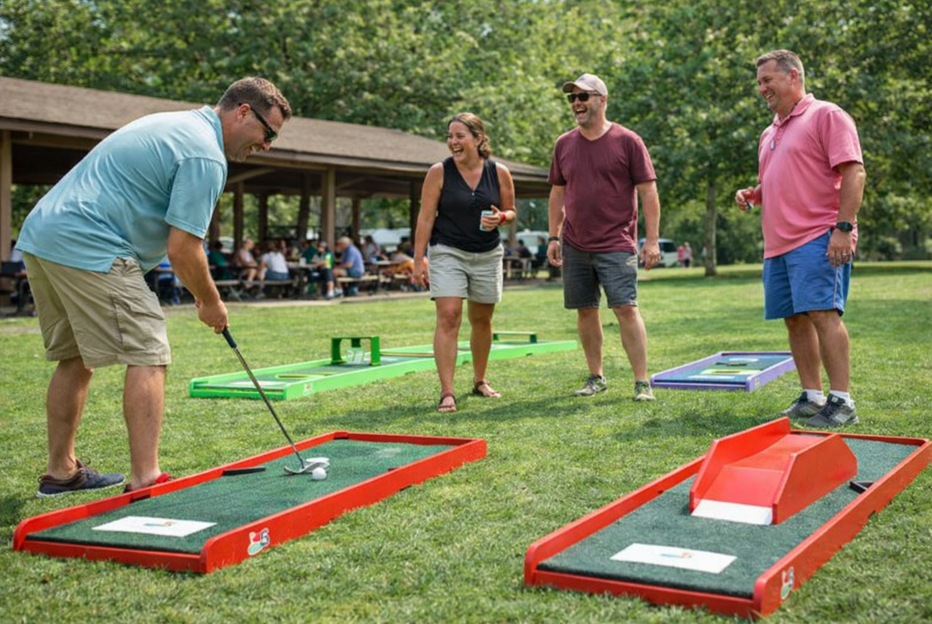 Portable mini golf rental setup at a company picnic in Greer, SC with adults laughing and playing on a sunny day in a park.