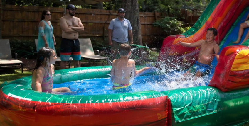 Children playing on a tropical-themed inflatable bounce house water slide rental at a backyard party in Greenville, SC. Realistic summer scene with kids splashing in the pool, sliding, and bouncing while adults supervise, perfect for last-minute May and early summer party rentals in Greenville.