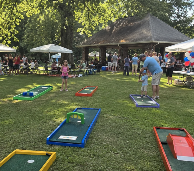 Portable mini golf rentals at a South Carolina family festival, fundraiser, or private event, with colorful lanes set up on a grassy lawn surrounded by guests, tents, tables, and a pavilion. Children play mini golf while adults socialize in a lively outdoor community setting.