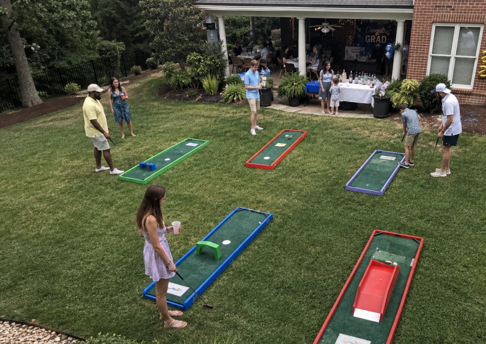 Portable mini golf rentals at a South Carolina graduation party, with colorful putting lanes arranged across a landscaped backyard. Guests play and mingle near a brick home, graduation decorations, tables, and balloons, creating a fun activity for the celebration.