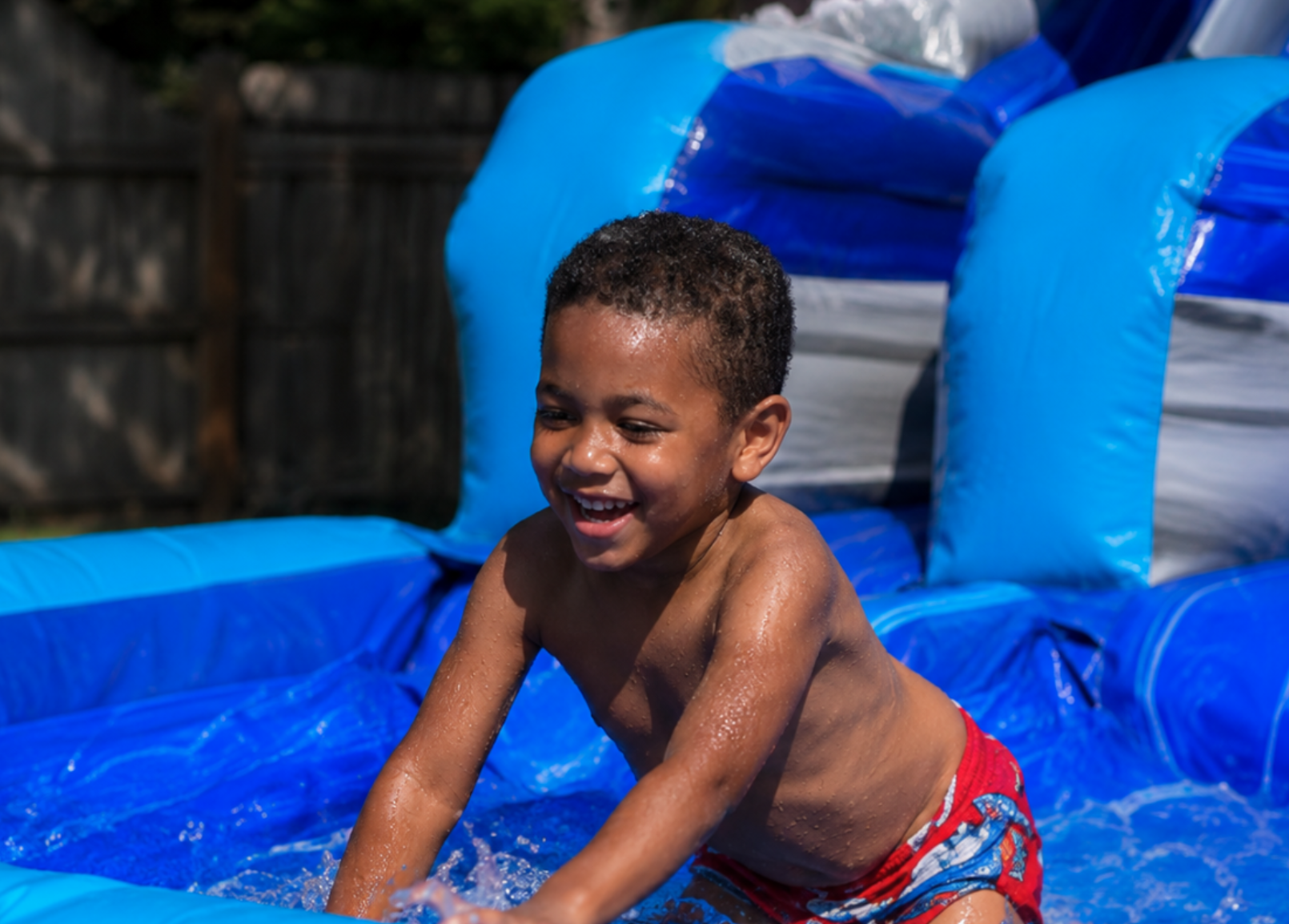 Smiling boy splashing and playing in the shallow pool of a blue inflatable water slide.