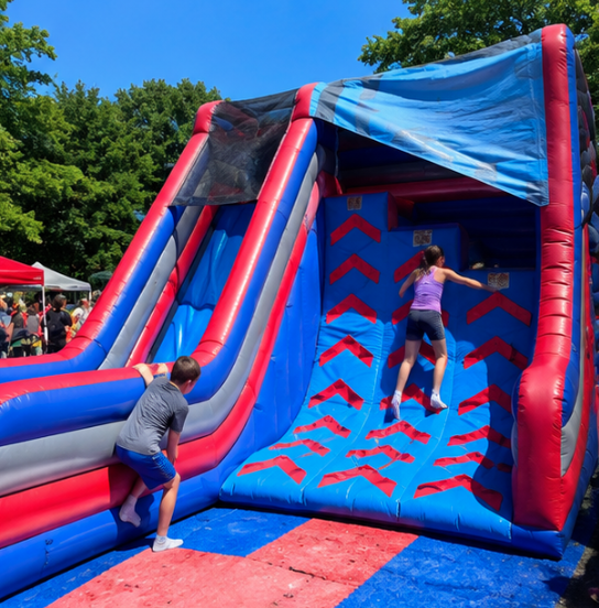 Kids climbing and playing on a blue and red inflatable ninja wall at an outdoor community event.