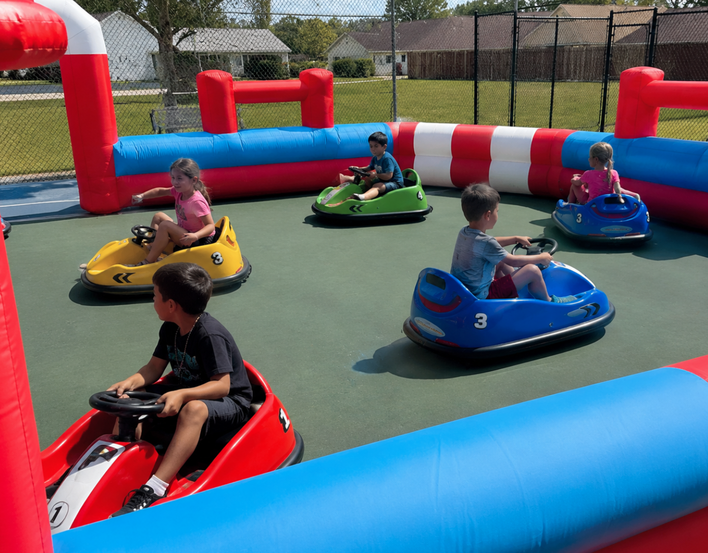 Children riding bumper cars on a smooth play surface inside a red, blue, and white inflatable arena.