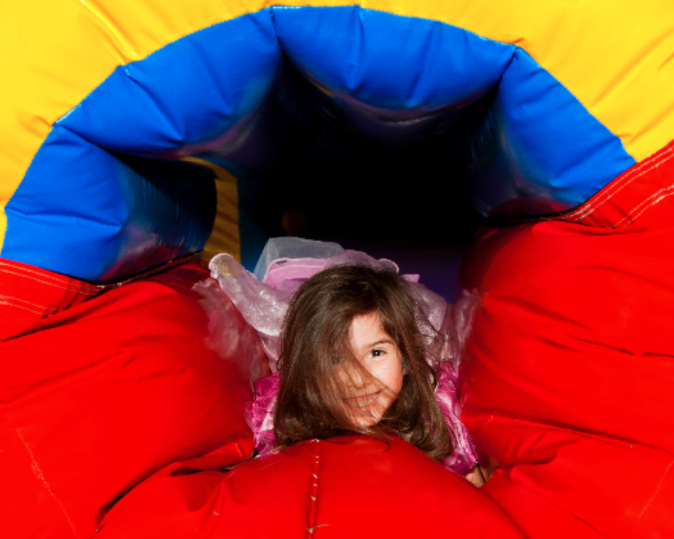 Young girl in a pink outfit smiling while playing between red inflatable walls inside a colorful bounce house.