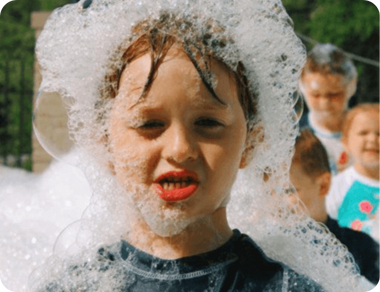 Child covered in white foam, smiling during an outdoor bubble or foam party.