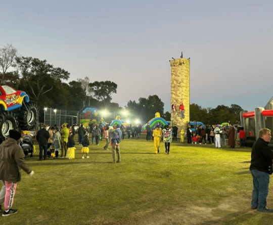 Outdoor event at dusk with people gathered around inflatable attractions, a rock climbing wall, and monster trucks.