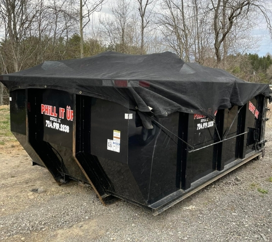 PHiLL It Up Disposal, LLC black dumpster with a tarp cover placed on gravel near tree