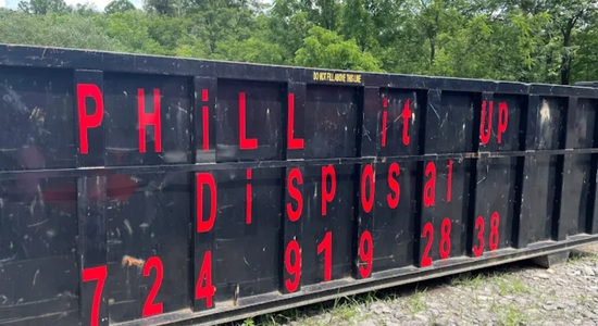 Large black PHiLL It Up Disposal, LLC roll-off dumpster with red lettering on a gravel surface near trees