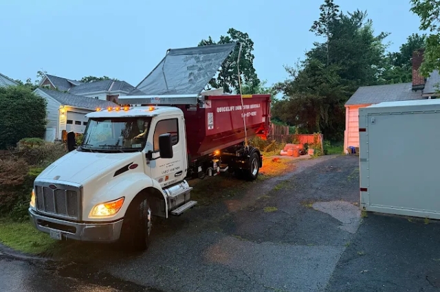 At dusk on a wet road, a roll-off truck completes a pickup for QuickLift Dumpster Rental