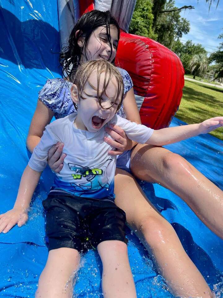 A joyful moment captured as two children, with expressions of sheer delight, slide down a wet, blue inflatable water slide in Ocala, FL, provided by Rebound Party Rentals. The foreground shows a young boy with water splashing around him and a girl behind him, both enjoying a sunny day of outdoor fun.