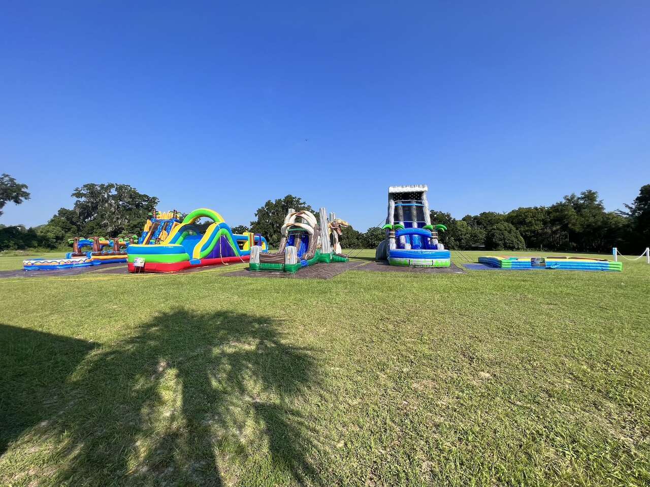An expansive outdoor setup of colorful inflatable bounce houses and water slides by Rebound Party Rentals in a green park in Ocala, FL. The scene includes a tropical-themed slide, a multi-colored castle bounce house, and a large blue water slide under a clear blue sky, perfect for summer festivities and family fun days.