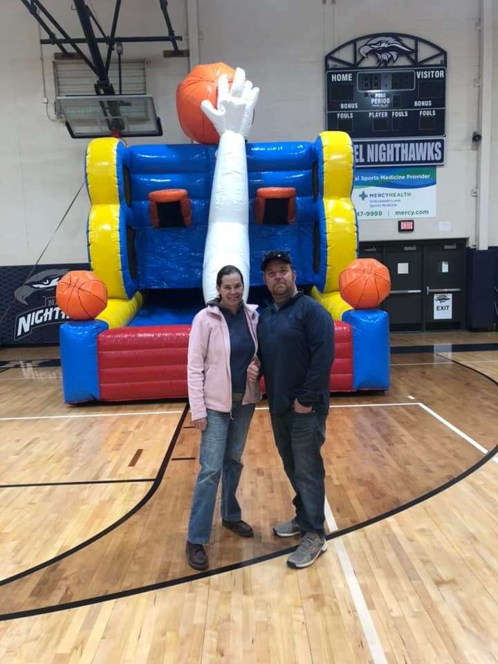 Ian and Lynette Mathieu, the husband and wife team behind Party Go Round, standing proudly in front of a colorful bounce house shaped like a giant basketball hoop, available for rent in Cincinnati.